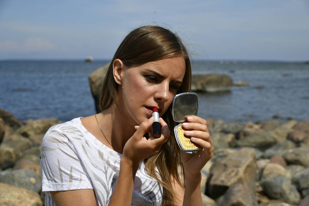 A focused young woman applying red lipstick while looking into a compact mirror on the scenic rocky shores of Muraste, Estonia.