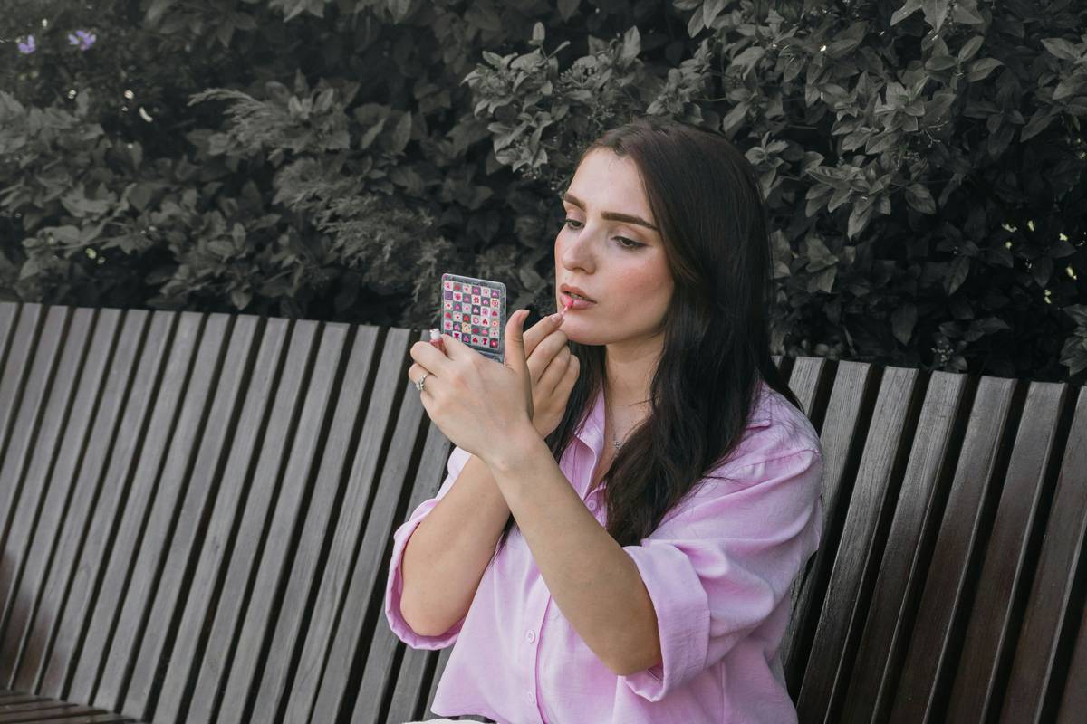A woman in a pink shirt applying lip gloss using a small patterned mirror